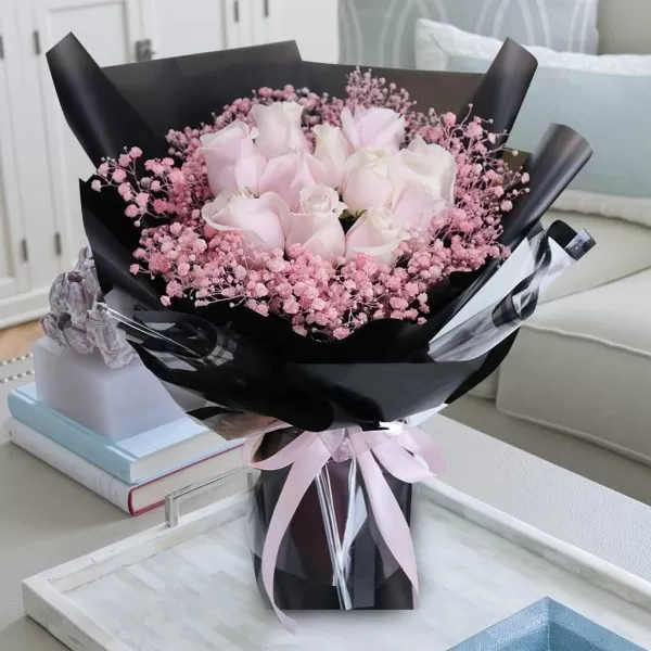 A close-up shot of a bouquet featuring pink roses and small pink baby's breath flowers, wrapped in black paper and tied with a light pink ribbon, placed on a light-colored surface with decorative items and a blurred background.