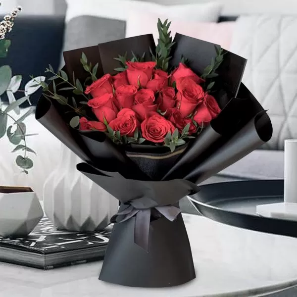 A bouquet of red roses with green leaves, wrapped in black paper and tied with a gray ribbon, placed on a light-colored surface with a blurred domestic background.