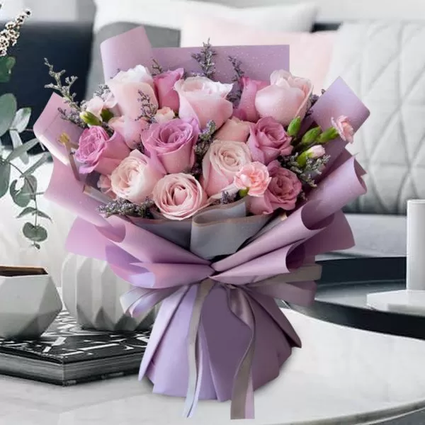 A close-up shot of a bouquet featuring light pink roses, deep pink roses, and small pink carnations, with small purple flowers and greenery, wrapped in light purple paper and tied with a light purple ribbon, placed on a light-colored surface with decorative items and a blurred background.
