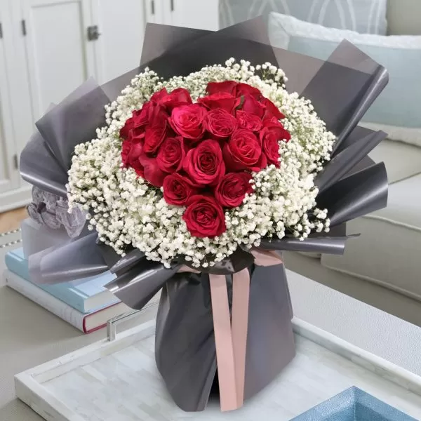A bouquet of red roses arranged in a heart shape, surrounded by white baby's breath flowers, wrapped in gray paper and tied with a pink ribbon, placed on a gray tray on a table with books and a sofa in the background.