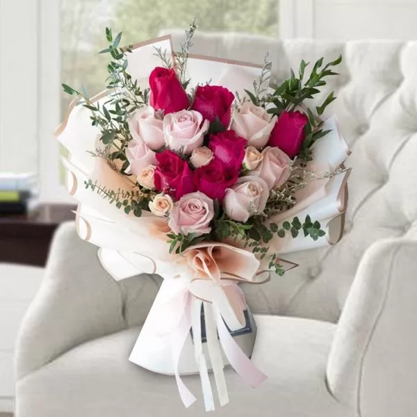 A bouquet of pink and red roses with green eucalyptus leaves, wrapped in white paper and tied with a pink ribbon, placed on a white armchair with a blurred background of a room with a window.