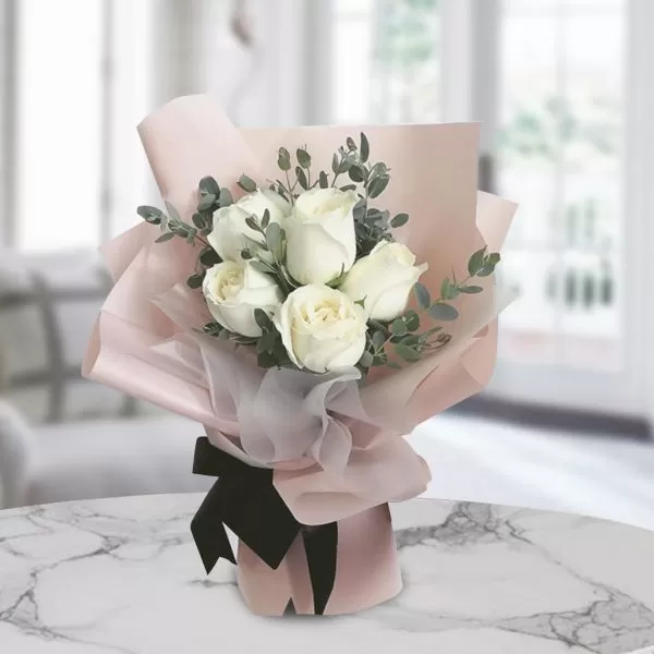 A bouquet of white roses with green eucalyptus leaves, wrapped in light pink paper and tied with a black ribbon, placed on a marble table with a blurred background.