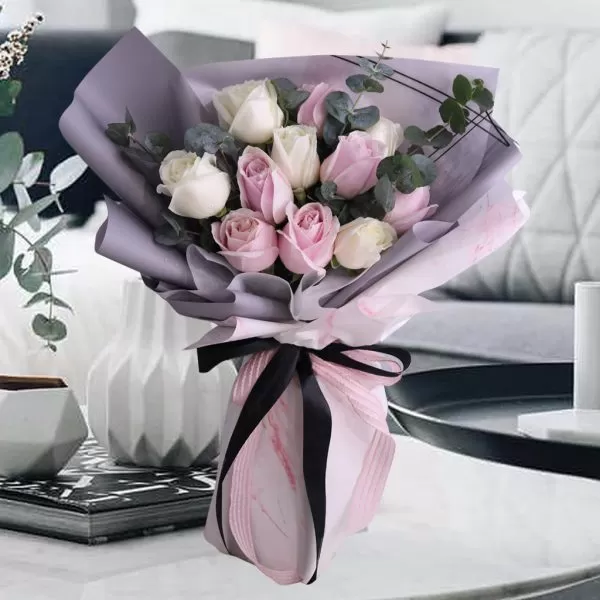 A close-up shot of a bouquet featuring pink and white roses, with eucalyptus leaves, wrapped in a combination of light purple and pink paper, tied with a black ribbon, placed on a light-colored surface with decorative items and a blurred background. A close-up shot of a bouquet featuring pink and white roses, with eucalyptus leaves, wrapped in a combination of light purple and pink paper, tied with a black ribbon, placed on a light-colored surface with decorative items and a blurred background.