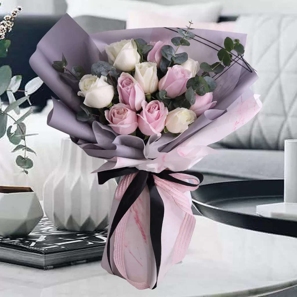 Lara A close-up shot of a bouquet featuring pink and white roses, with eucalyptus leaves, wrapped in a combination of light purple and pink paper, tied with a black ribbon, placed on a light-colored surface with decorative items and a blurred background. A close-up shot of a bouquet featuring pink and white roses, with eucalyptus leaves, wrapped in a combination of light purple and pink paper, tied with a black ribbon, placed on a light-colored surface with decorative items and a blurred background.