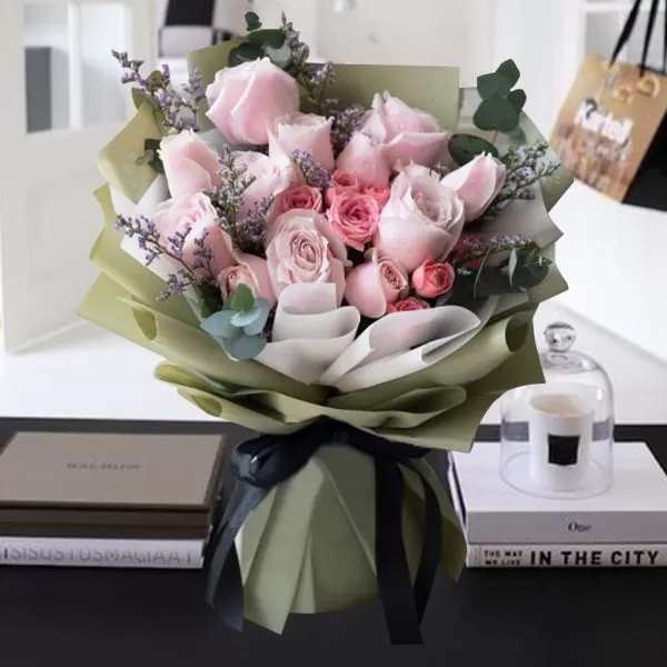 A close-up shot of a bouquet featuring light pink roses, smaller pink roses, and small purple flowers, arranged with greenery, wrapped in light green paper and tied with a black ribbon, placed on a table with two books, one titled "The Way We Live In The City," and a glass cloche with a white candle.