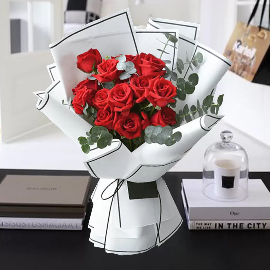 Rory A bouquet of red roses with green eucalyptus leaves, wrapped in white paper with black lines and tied with a black ribbon, placed on a table with two books and a glass cloche with a candle inside. The background is blurred, showing a room with a window and a shopping bag.