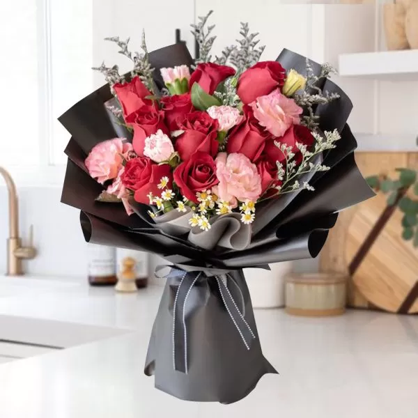 This image shows a bouquet of red and pink roses, pink carnations, and small white daisies, wrapped in black paper and tied with a gray ribbon, placed on a white countertop with a blurred kitchen background.