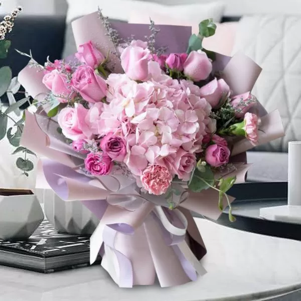 A close-up shot of a bouquet featuring light pink roses, deep pink roses, a light pink hydrangea, small pink carnations, and small purple flowers, with eucalyptus leaves, wrapped in light purple paper and tied with a light purple ribbon, placed on a light-colored surface with decorative items and a blurred background.