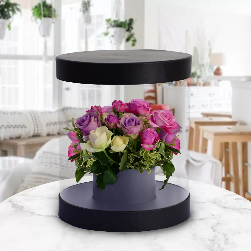 A round, clear flower box with a black base and lid, containing a bouquet of pink and purple roses and green leaves, placed on a marble table with a blurred background of a living room. The box appears to be floating due to the reflection on the table.