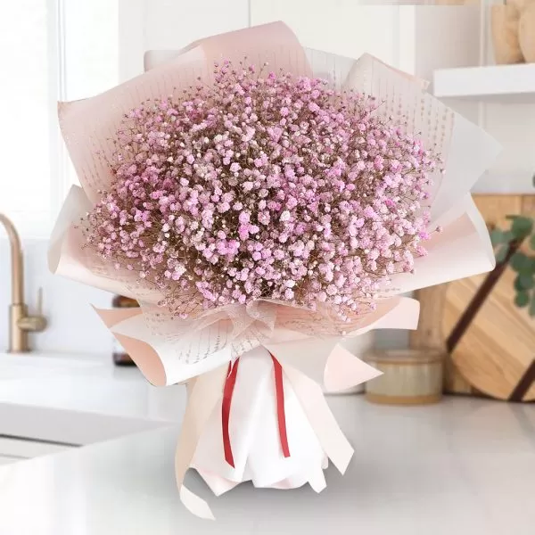 A close-up shot of a bouquet featuring a dense cluster of small pink baby's breath flowers, wrapped in pink and white paper and tied with a light pink ribbon, placed on a white countertop with a blurred kitchen background.