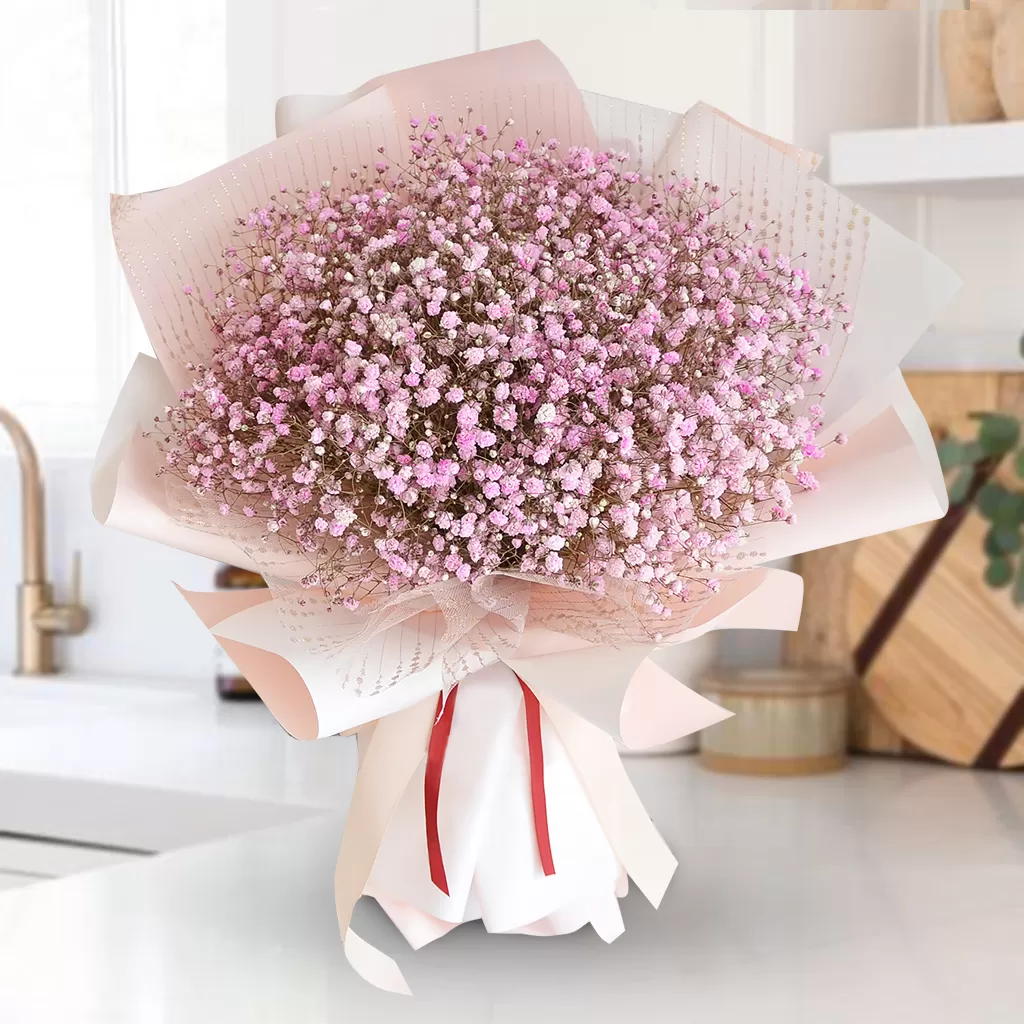 A close-up shot of a bouquet featuring a dense cluster of small pink baby's breath flowers, wrapped in pink and white paper and tied with a light pink ribbon, placed on a white countertop with a blurred kitchen background.