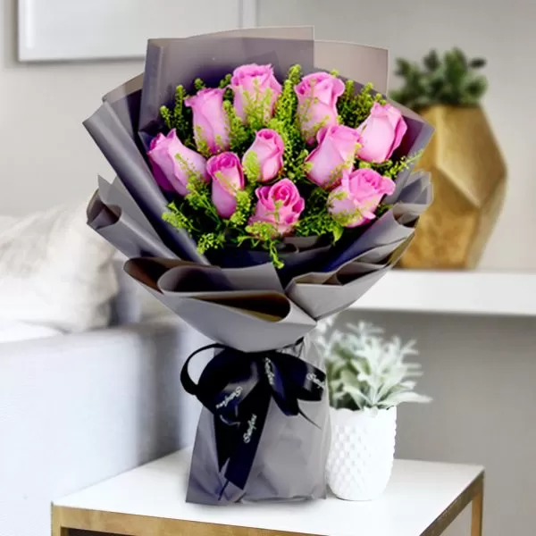 A close-up shot of a bouquet featuring pink roses with green filler flowers, wrapped in gray paper and tied with a black ribbon, placed on a light-colored surface with a blurred background showing a gold vase and a small plant.