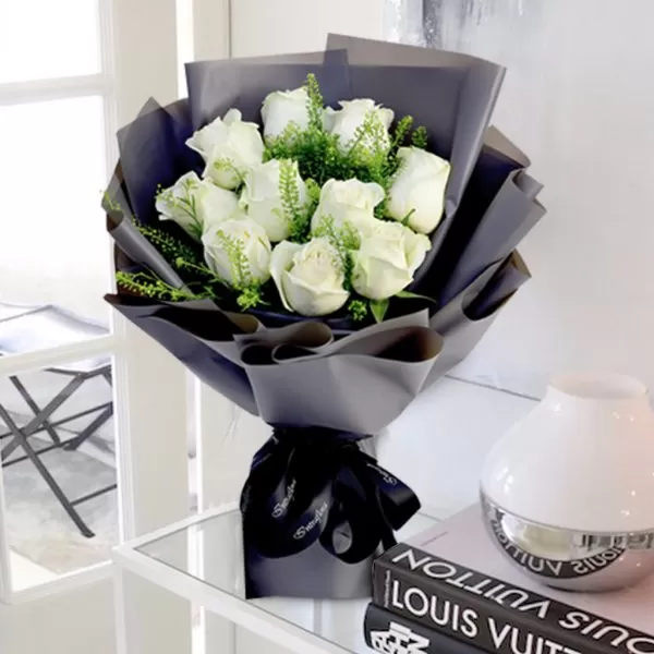 A bouquet of white roses with green filler flowers, wrapped in dark gray paper and tied with a black ribbon, placed on a white table with two books and a white vase. The background is blurred, showing a room with a window.