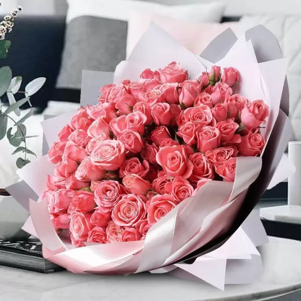 A bouquet of pink roses, wrapped in light gray paper and tied with a pink ribbon, placed on a table with books and decorative items, with a blurred background of a living room. The bouquet appears to be made up entirely of roses.