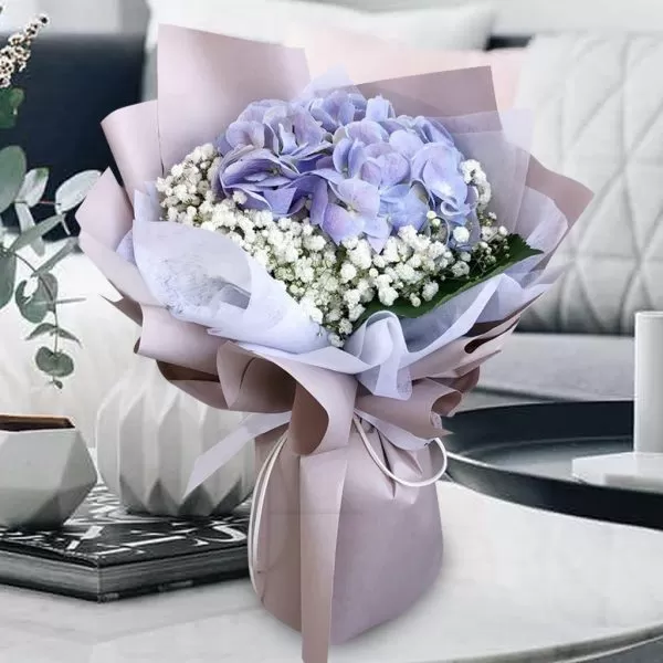 A close-up shot of a bouquet featuring purple hydrangeas and small white baby's breath flowers, wrapped in layers of light purple paper, placed on a light-colored surface with decorative items and a blurred background.