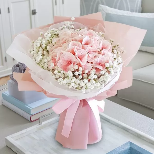 A close-up shot of a bouquet featuring pink hydrangeas and small white baby's breath flowers, wrapped in layers of pink and white paper, tied with a pink ribbon, placed on a light-colored surface with decorative items and a blurred background.