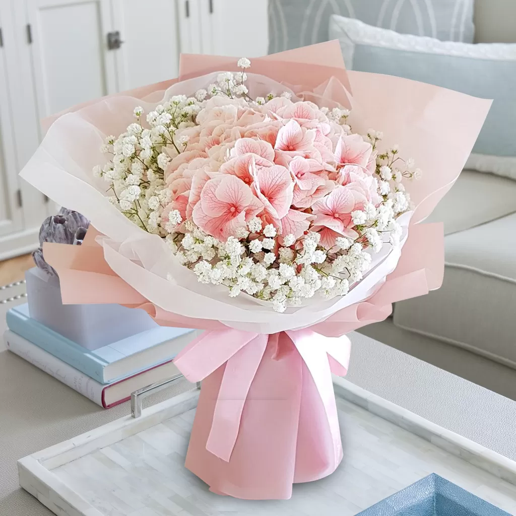 A close-up shot of a bouquet featuring pink hydrangeas and small white baby's breath flowers, wrapped in layers of pink and white paper, tied with a pink ribbon, placed on a light-colored surface with decorative items and a blurred background.