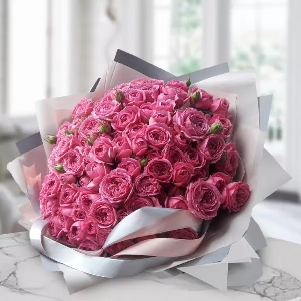 A bouquet of pink roses, wrapped in light gray paper and tied with a silver ribbon, placed on a marble table with a blurred background. The bouquet appears to be made up entirely of roses.