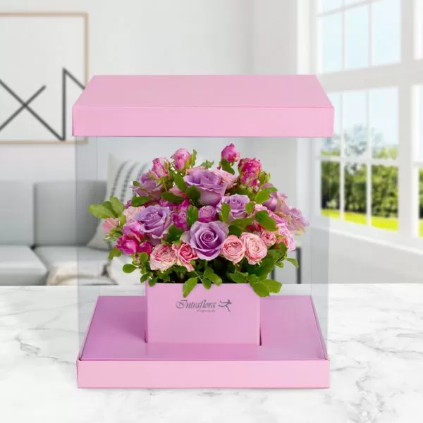 A square, clear flower box with a pink base and lid, containing a bouquet of pink and purple roses and green leaves, placed on a marble table with a blurred background. The box also features the "Intraflora" logo. The box appears to be floating due to the reflection on the table.