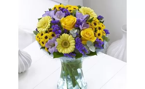 A glass vase filled with yellow roses, yellow gerbera daisies, purple iris flowers, and small yellow flowers, placed on a white table.