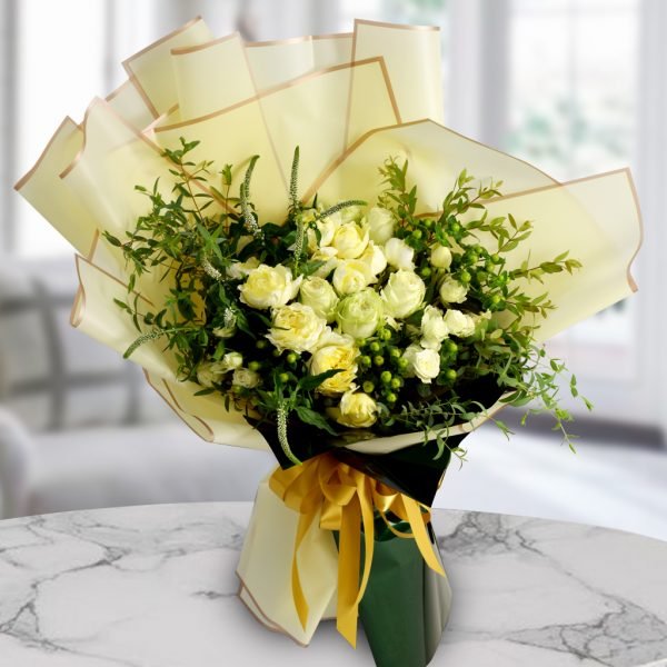 A bouquet of yellow and white roses with green filler flowers, wrapped in light yellow paper and tied with a yellow ribbon, placed on a marble table with a blurred background. The background shows a room with a chair.