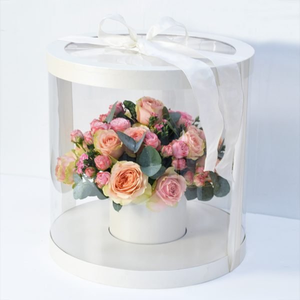 A round, transparent flower box with a white base and lid, tied with a white ribbon, containing a bouquet of pink and peach roses and greenery.