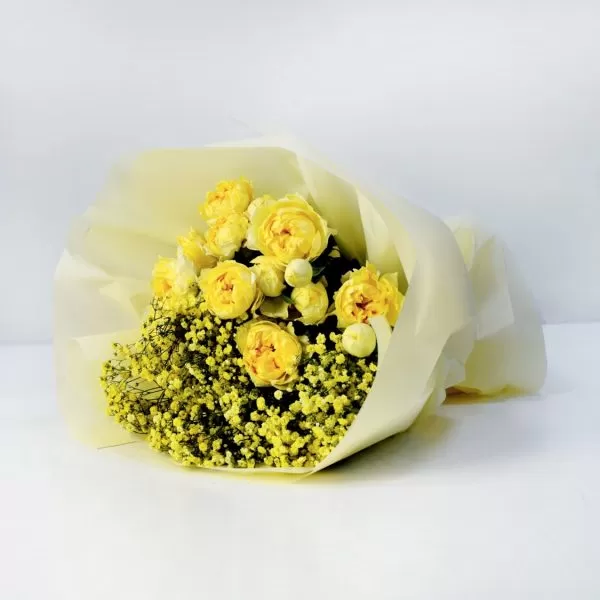 A bouquet of yellow roses and yellow baby's breath flowers, wrapped in light yellow paper, against a light background.