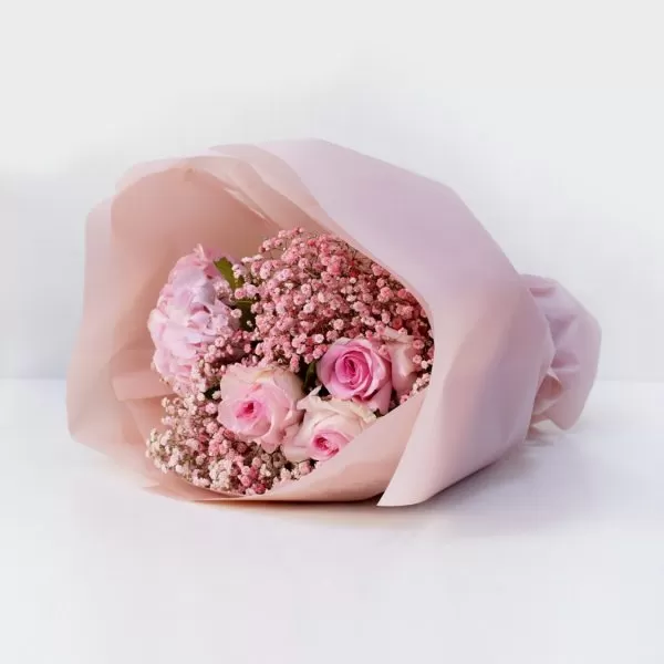 A close-up shot of a bouquet featuring pink roses and small pink baby's breath flowers, wrapped in light pink paper, placed on a white surface.