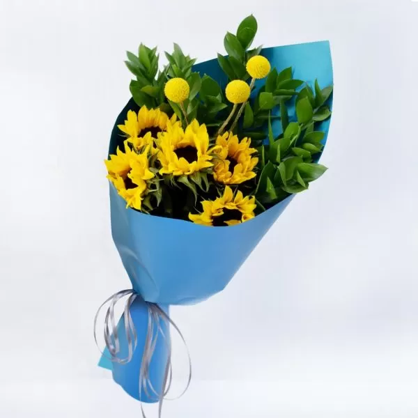 A close-up shot of a bouquet featuring yellow sunflowers, yellow billy balls, and green leaves, wrapped in bright blue paper and tied with a gray ribbon.