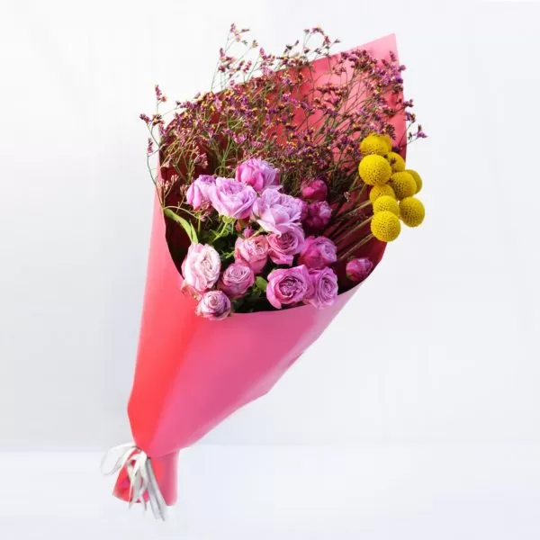 A bouquet of pink roses, purple dried flowers, and yellow billy balls wrapped in pink paper, tied with a silver ribbon, against a light background.