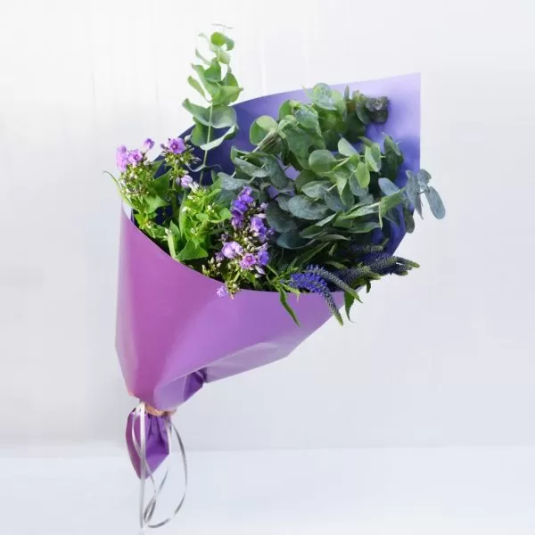A bouquet of purple flowers and eucalyptus leaves wrapped in purple paper, tied with a silver ribbon, against a light background.