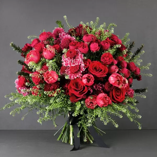 A lush bouquet of red roses, red tulips, red carnations, and green filler flowers, tied with a black ribbon, against a dark gray background.
