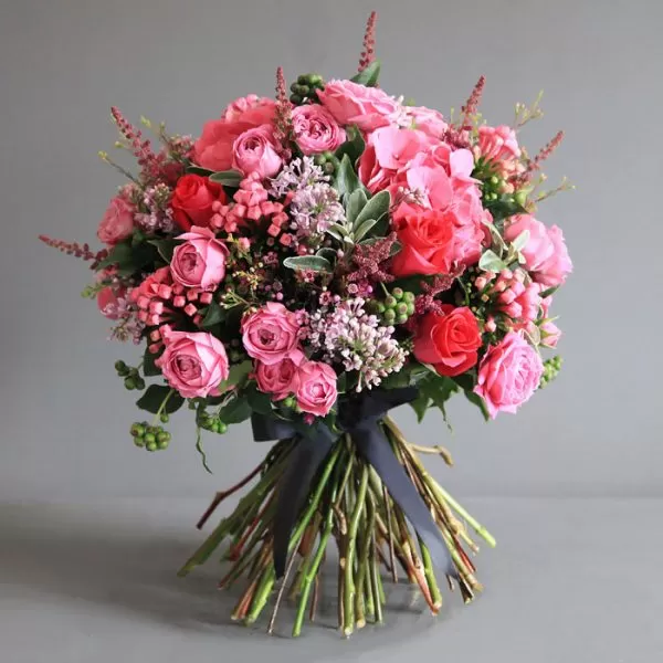 A bouquet of pink roses, pink hydrangeas, pink spray roses, purple waxflowers, green berries, and green filler flowers, tied with a black ribbon, against a dark gray background.