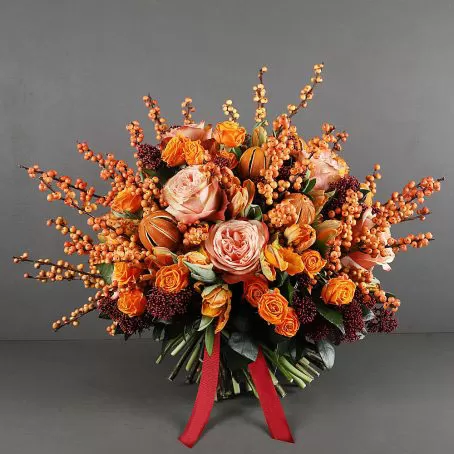 A bouquet of orange roses, orange ranunculus, orange berries, and burgundy filler flowers, tied with a red ribbon, against a dark gray background.