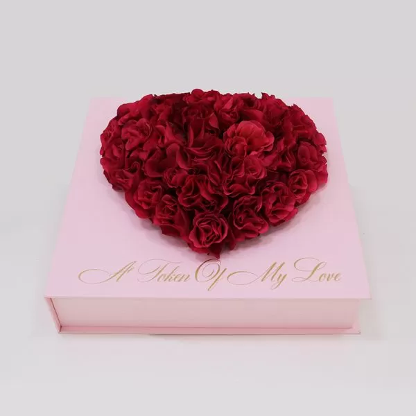 A light pink square box with a heart-shaped arrangement of dark red flowers on top. The phrase "A Token Of My Love" is written in gold script on the front of the box.