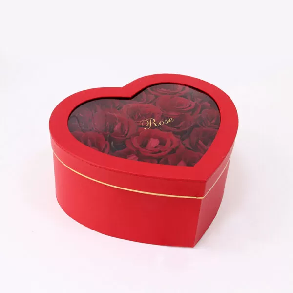 A red, heart-shaped box with a transparent lid, showcasing red roses inside. The word "Rose" is written in gold on the transparent lid.