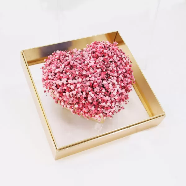 A heart-shaped arrangement of small pink and white flowers inside a square, gold-edged frame with a transparent base.