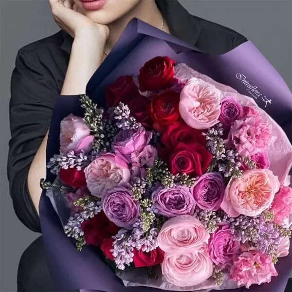 A close-up shot of a bouquet featuring red, pink, and purple roses, pink carnations, and small purple flowers, wrapped in dark purple paper with the "Intraflora" logo, held by a person with dark hair, wearing a black blazer, with only their face and shoulder visible.