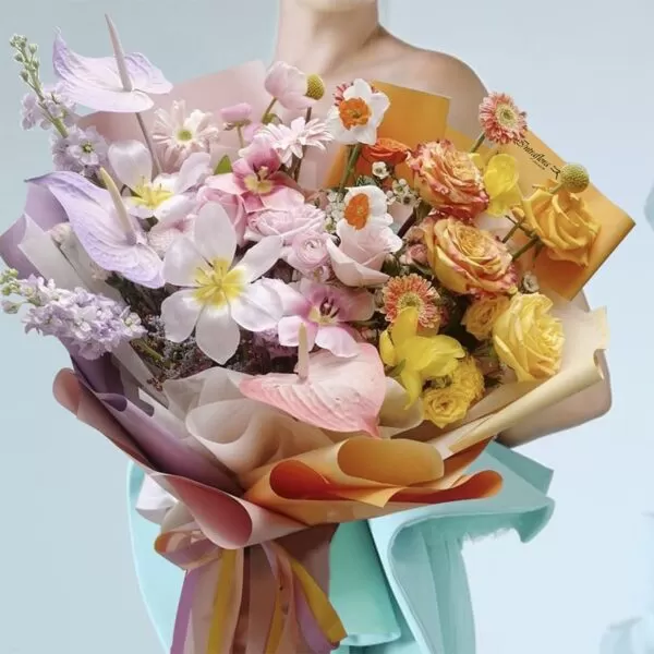 A person in a light blue dress holding a bouquet of light pink tulips, light pink roses, yellow roses, orange ranunculus, yellow gerbera daisies, purple anthuriums, and white snapdragons, wrapped in light purple and orange paper. The bouquet also features a yellow card with the word "Intraflora" on it.