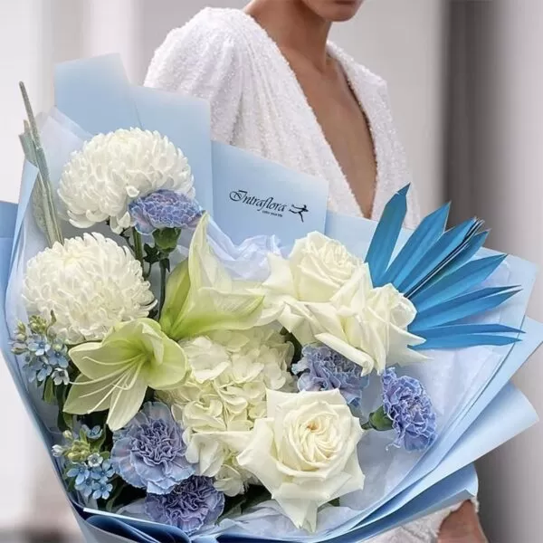 A woman in a white dress holding a bouquet of white chrysanthemums, white roses, white lilies, blue carnations, blue delphiniums, and blue palm leaves, wrapped in light blue paper. The bouquet also features the "Intraflora" logo.