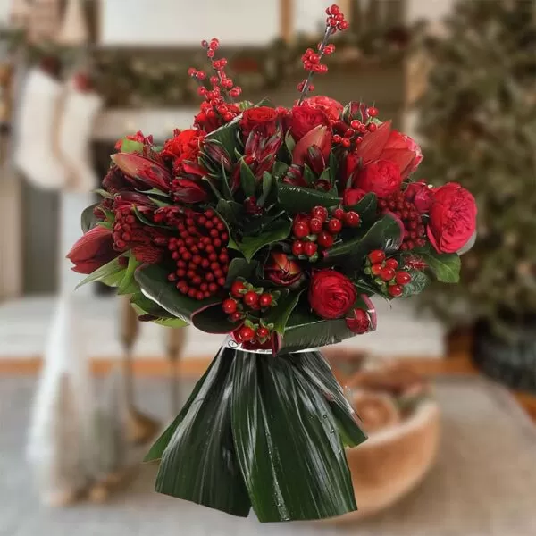 This image shows a festive bouquet of red flowers and berries, including roses, tulips, and hypericum berries, wrapped in green leaves and placed in a blurred Christmas-themed setting. The background suggests a cozy home with decorations like stockings and a Christmas tree.