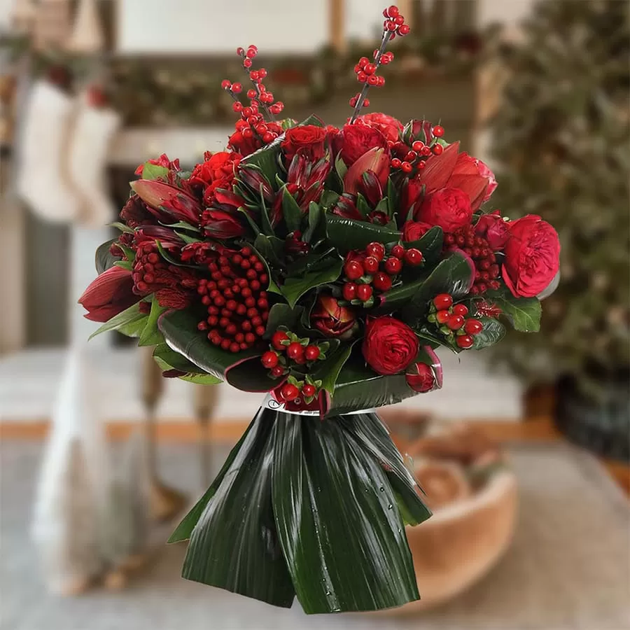 ROSEBERRRIES This image shows a festive bouquet of red flowers and berries, including roses, tulips, and hypericum berries, wrapped in green leaves and placed in a blurred Christmas-themed setting. The background suggests a cozy home with decorations like stockings and a Christmas tree.