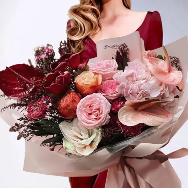 A woman in a red dress holding a bouquet of pink and orange roses, pink hydrangeas, pink anthuriums, burgundy calla lilies, burgundy carnations, and burgundy amaranthus, wrapped in light brown paper. The bouquet also features the "Intraflora" logo.