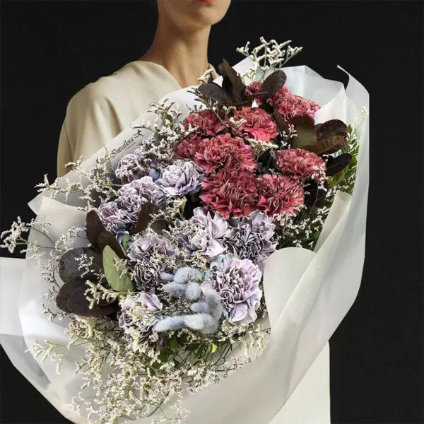 A person in a white dress holding a bouquet of purple and burgundy carnations, white statice, and dark purple leaves, wrapped in white paper. The bouquet also features a small blue tag with the word "Intraflora" on it.