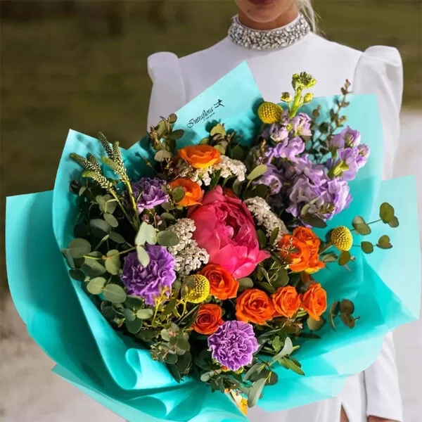A woman in a white dress with a jeweled neckline holding a bouquet of pink peonies, orange roses, yellow craspedia, purple carnations, purple delphiniums, and green eucalyptus leaves, wrapped in light blue paper. The bouquet also features the "Intraflora" logo.