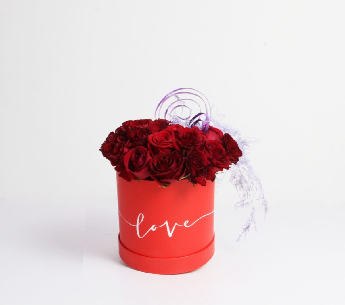 A red, cylindrical flower box filled with dark red roses, with the word "love" written in white script on the side. The box also features purple decorative twigs and light purple feathery decorations, against a light background.