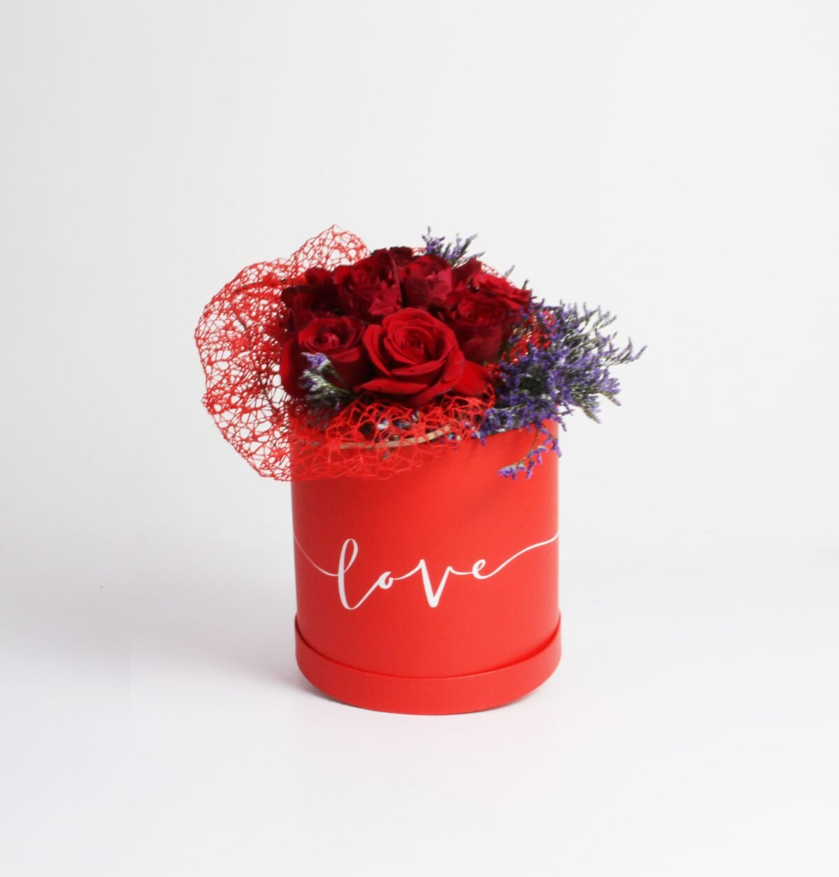 A red, cylindrical flower box filled with dark red roses and purple waxflowers, with the word "love" written in white script on the side. The box also features a red mesh decoration, against a light background.