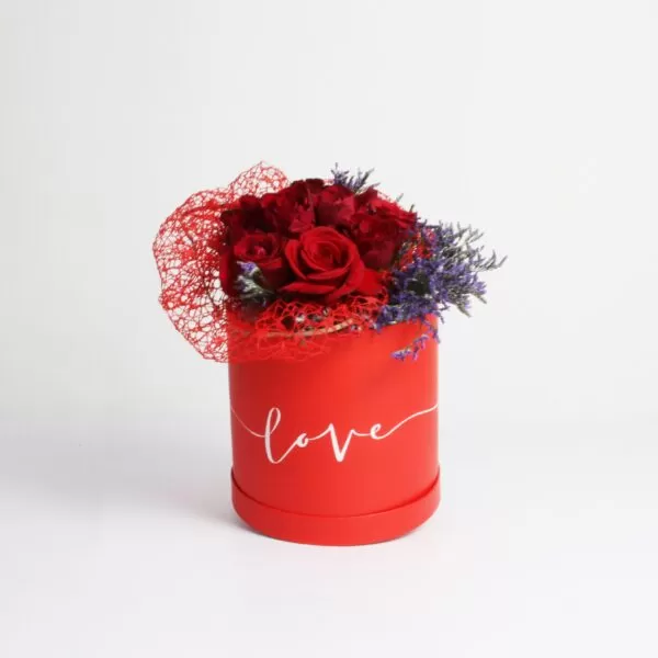 A red, cylindrical flower box filled with dark red roses and purple waxflowers, with the word "love" written in white script on the side. The box also features a red mesh decoration, against a light background.