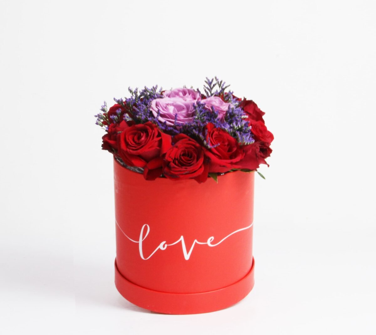 A red, cylindrical flower box filled with dark red roses, purple roses, and purple waxflowers, with the word "love" written in white script on the side, against a light background.