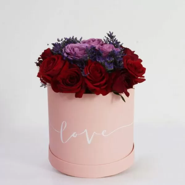 A pink, cylindrical flower box filled with dark red roses, purple roses, and purple waxflowers, with the word "love" written in white script on the side, against a light background.